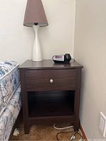 Bedside table with drawer and cubby, small lamp on top, and Audiovox clock radio beside the lamp