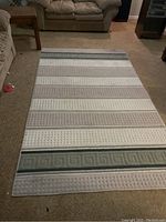 Full view of the large carpet laid out on floor, showing front pattern with stripes, textured details, and Greek key band on one end. Light neutral colors of gray, white, and beige.