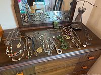 Overview of multiple costume jewelry necklaces laid out on dark wood dresser with mirror. Necklace holder on right side includes a stand and bowl with rings.