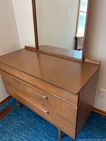 Image showing front view of mid-tone brown wood and press board dresser with three drawers, integrated wooden handles, tapered legs, and attached rectangular mirror in wooden frame.