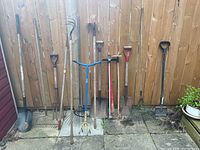 Photo showing 11 assorted gardening tools leaning against a wooden fence on a concrete surface. Tools include shovels, rakes, pitchfork, hoe, manual soil aerator, and hand cultivator, all used but working.