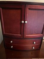 Jewelry box closed showing mahogany wood finish with two top doors and four drawers below, all with round metal knobs.