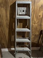 Front view of a metal 5-step ladder leaning against wood panel wall.