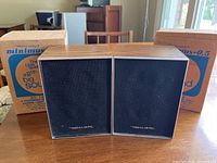 Front view of two Realistic Minimus 0.5 speakers side by side with original cardboard boxes behind them on a wooden table.