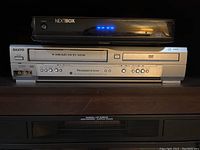 Photo showing the front of a silver Sanyo 4-Head Hi-Fi VCR and DVD player combo unit with a black Nextbox device above it.
