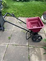Full side view of the seed and fertilizer spreader showing red plastic hopper, black frame, wheels, and handlebar with control trigger.