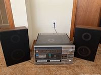 Photo showing the full stereo system with two black speakers on either side, power cord plugged in the wall outlet, and device placed on carpeted floor.
