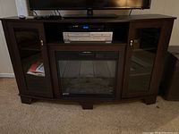 Front view of dark wood TV console with electric fireplace insert center and glass door cabinets on each side, TV and stereo electronics placed on top and in open shelf but not included in lot.