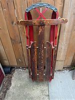 Full front view of vintage sled showing wooden slats, red runners, metal handle and attached yellow rope.