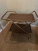Full view of vintage tea cart showing two brown trays with metal frame and handles, and rolling ball feet on carpeted floor.