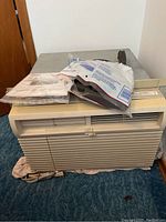 Front view of window air conditioner showing beige grille and controls, with manual and accessories on top.