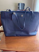 Front view of navy blue Tory Burch purse with logo and handles visible, placed on wooden surface.