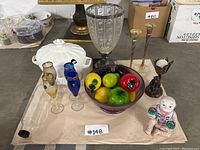 Wide shot showing entire assortment on beige cloth including glasses, dishes, bowl with glass fruit, candlesticks, and figurines.