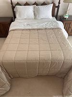 Full view of beige quilted comforter laid on bed with three white pillows in white pillowcases behind it.