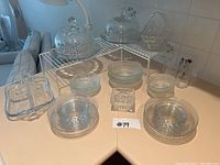 Overview of full lot showing crystal cake stands on top of white metal shelf risers alongside various clear glass plates, bowls, and platters arranged on table.