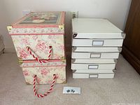 Two stacked floral-patterned storage boxes with red and white rope handles next to four stacked white organizational boxes with metal corners and label holders on a carpeted floor.