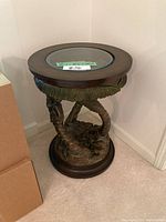 Round wood and glass side table with sculpted base shaped like intertwined palm tree trunks and leaves, placed next to cardboard boxes in living room corner.
