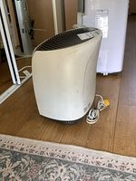 Side view of white Enviiracaire air purifier showing curved design and power cord on wood floor near carpet edge.