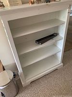 Full view of white painted wood bookcase with four shelves, empty except for a small boxed item on one shelf.