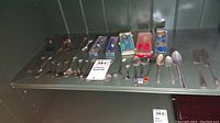 Shelf display with more than twenty silver plate and stainless steel collector spoons. Some in plastic cases, some loose. Various sizes and decorative details on handles.