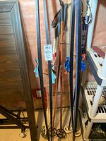 Cross country skis (black and white pairs) and four ski poles leaning against the wall with tags attached.
