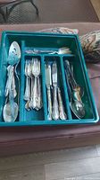 Mismatched cutlery arranged in a green plastic tray showing various types of spoons, forks, cutters, and serving utensils with ornate handles.