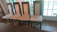 Four wood dining chairs with beige upholstered seats and backs aligned in front of a window.