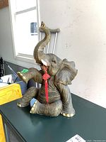 Full view of resin sitting elephant statuette showing raised trunk, red tassel, and textured skin details.