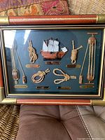 Framed nautical art showing a wood sailing ship, various rope knots, and wooden plaques with knot names on a dark green background in a frame with leather corners and brass accents.