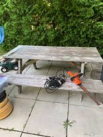 Photo showing weathered wooden picnic table on patio with hedge trimmer on bench.