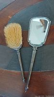 Photo of handheld mirror and hairbrush with silver handles and natural bristles, lying side by side on a wood surface.