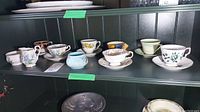 Shelf with assorted fine china teacups and saucers, cream and sugar bowls displayed on green wooden shelf with green tape marks.