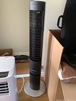 Full view of the black and gray Kaz Inc tower fan standing on a wooden floor, showing front grille and stable round base.