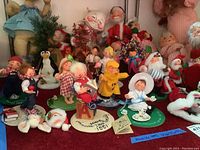 Photo showing multiple small Annalee dolls arranged on a red surface with bases, including dolls in overalls, checkered dress, yellow raincoat and sailor outfit, plus a penguin and some Santa dolls.
