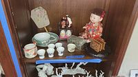 Full view of the collection including dolls, tea set, decorative eggs, fan, and small wooden box on dark wood shelf