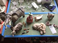 Photo of large gray ceramic elephants, sleeping beige pigs, Royal Doulton figurine, ceramic cow, red snake, and small animal figurines on green surface
