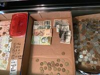 Photo shows a cardboard container holding foreign coins and various banknotes, some visible notes from Netherlands and Romania.