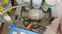 Full view of the Craftsman bench grinder with power cord and light, seen on a dusty floor surrounded by fertilizer containers in the background