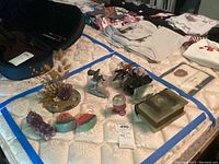 Overview of lot on mattress showing various polished stone art pieces including flower arrangement, watermelon slice stones, amethyst crystals, metal child figure on stone base, hinged stone book box, and dried flower base.