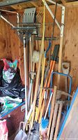 Various garden tools including different styles of rakes, shovels, and handle tools arranged against a wooden shed wall.