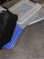 Four stackable storage containers in black, blue, gray, and clear with white lid featuring locking lids stacked on carpet.