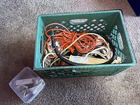 Green plastic crate containing several extension cords, ropes, 6-way plug outlets, lighting fixture tubes, fans, and more.