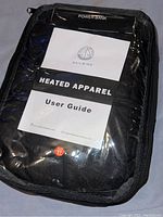 Photo of the heated apparel inside a transparent zippered case, showing the power bank compartment and Sailwind user guide inside the package.