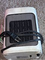 Top and side view of the white mini electric heater showing the black mesh grille and attached power cord with plug. Heater unit has some dirt and wear visible on the plastic housing.