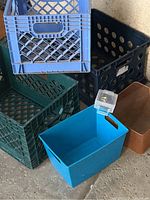 Photo showing six storage containers/crates in various colors and materials including plastic and metal, arranged on the ground against a wall.