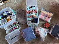 Photo showing sewing kits, storage containers, fabric bundles, pincushion, and assorted craft supplies arranged on carpeted floor.