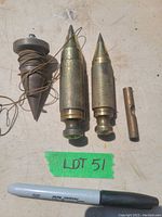 Four vintage plumb and level tools arranged on a surface near a marker pen, showing two brass knurled plumb bobs, one wooden top plumb bob with string, and a small brass level tool.