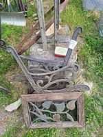 Photo showing two cast iron ornate bench ends and wooden framed decorative panel with leaf designs, placed on concrete and grass outdoors.