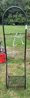 Full front view of the black metal freestanding coat rack with several horizontal bars for hooks and an arched top, standing outdoors on grass.