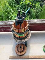 Full view showing whole fruit press with wooden basket, metal bands, central screw, handle, and separate wooden disc on the table